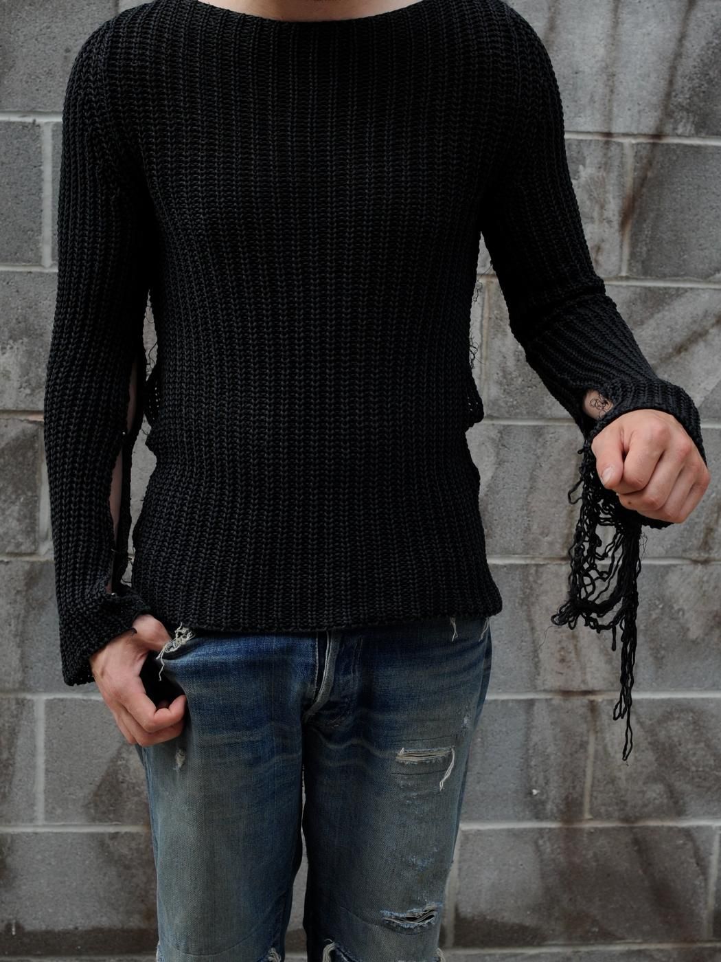 A black open knit sweater with a frayed cuff, worn against a grey block wall