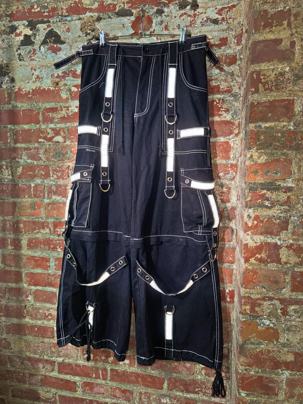 Black 3M Tripp NYC pants with reflective straps and D rings, hung against a painted brick wall
